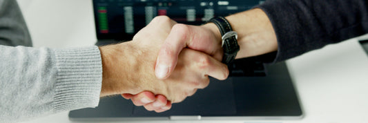 Two people shaking hands across a desk over a laptop showing price charts
